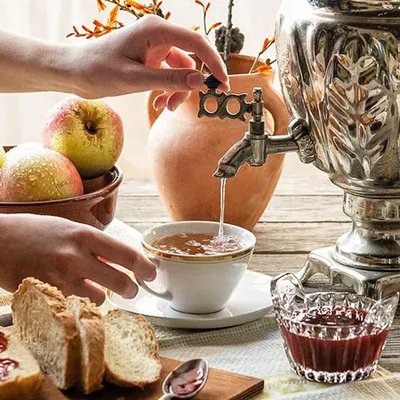 Dettaglio delle mani di una persona che prepara il tè usando un samovar metallico, con stoviglie e ingredienti ben visibili intorno.