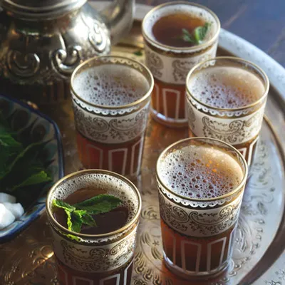 Gruppo di bicchieri pieni di infuso color ambra, serviti su un vassoio con decorazioni vegetali e luce soffusa.