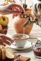 Dettaglio delle mani di una persona che prepara il tè usando un samovar metallico, con stoviglie e ingredienti ben visibili intorno.