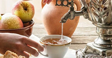 Dettaglio delle mani di una persona che prepara il tè usando un samovar metallico, con stoviglie e ingredienti ben visibili intorno.