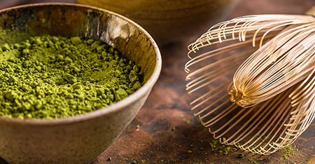 Ciotola con matcha verde brillante e frusta di bambù appoggiata accanto, fotografata in un set pulito e minimale.