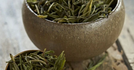 Primo piano di ciotola con tè verde essiccato, impiattato con cura su un tavolo o sfondo neutro, con ingredienti e decorazioni visibili attorno.